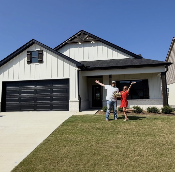A couple celebrating with Barbie in front of their new white farmhouse