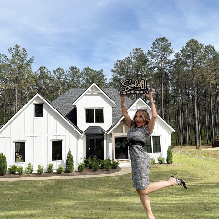 Barbie holding a Sold by @barbieperry sign in front of a white farmhouse